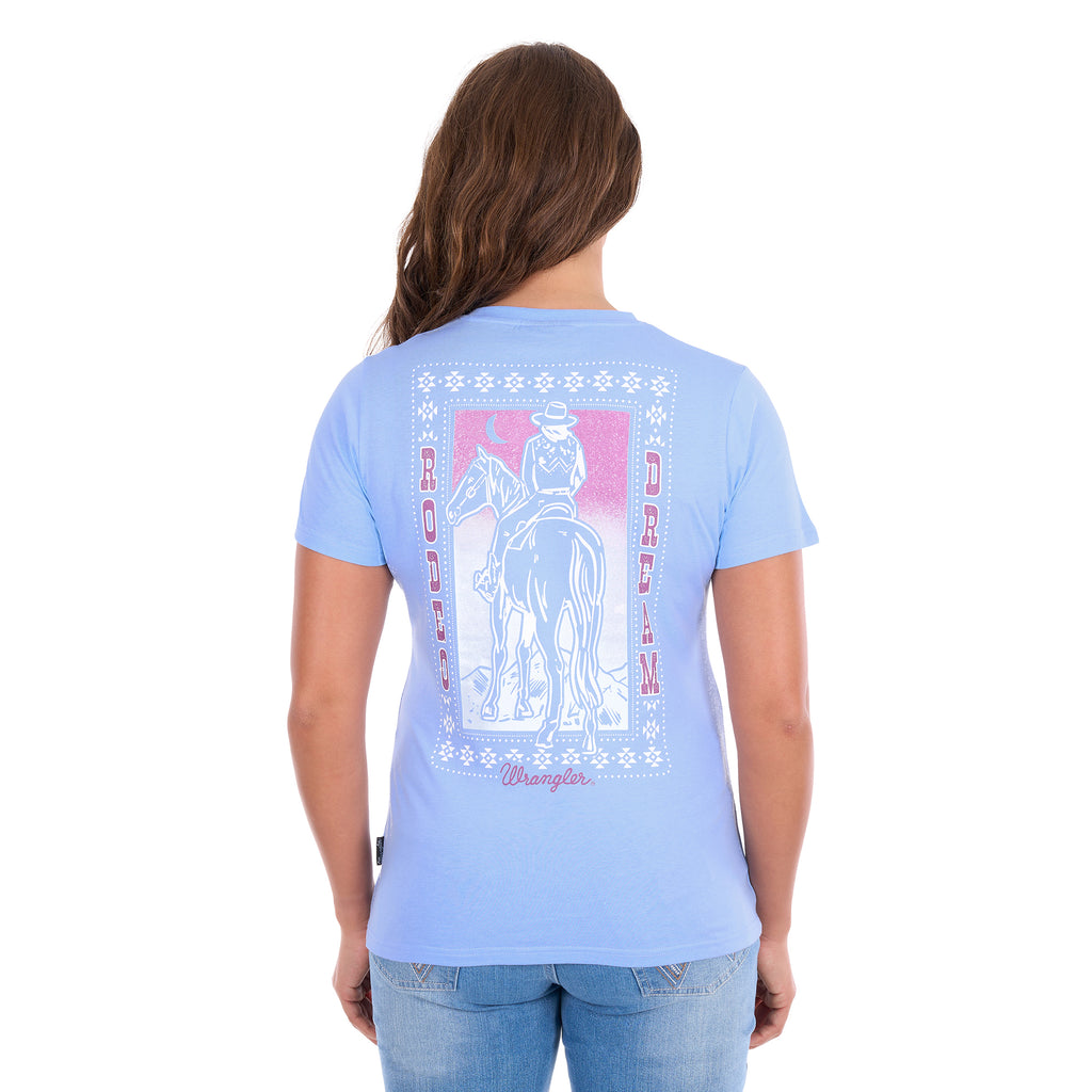 A woman with her back to the camera wearing a Wrangler Women's Lola Short Sleeved Tee (X6S2598642) in Light Blue featuring a large western 'Rodeo Dream' print in pink and white, with denim jeans.