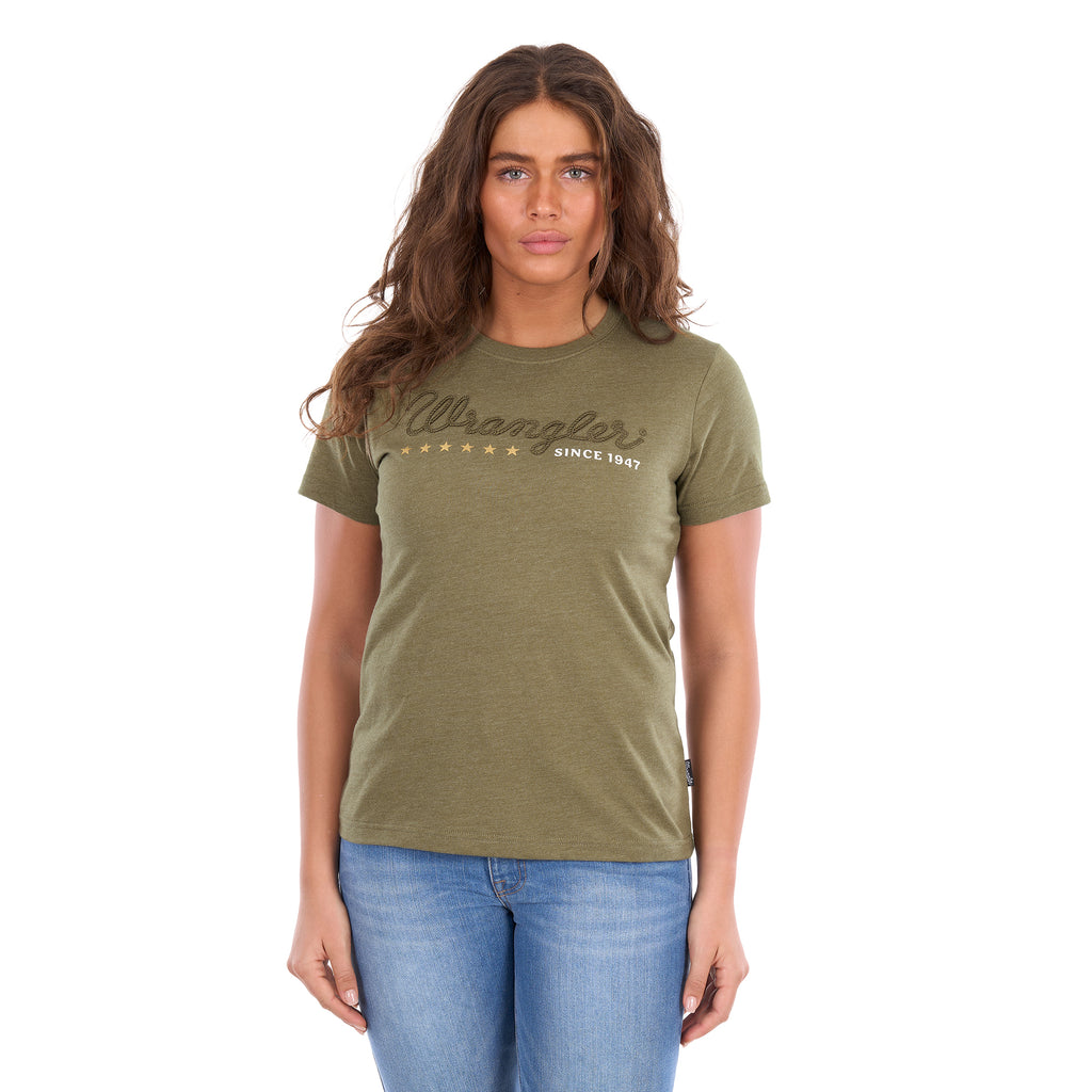 A woman modelling a Wrangler Women's Lucy Tee (XCP2598633) Khaki Marle with an embroidered script logo and gold stars on the chest, paired with blue jeans against a plain white background.