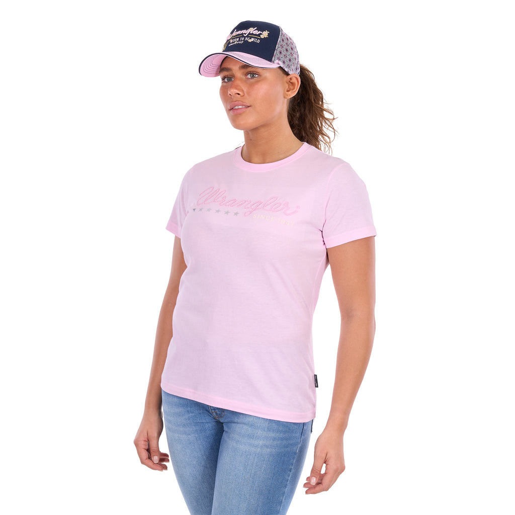 A woman modelling a pastel pink Wrangler Women's Lucy Short Sleeved T-Shirt (XCP2598633) with a pink script logo embroidered across the chest, paired with a navy and pastel pink Wrangler Bella High Profile Ponytail Trucker Cap (X6S2935CAP) and blue jeans against a plain white background.