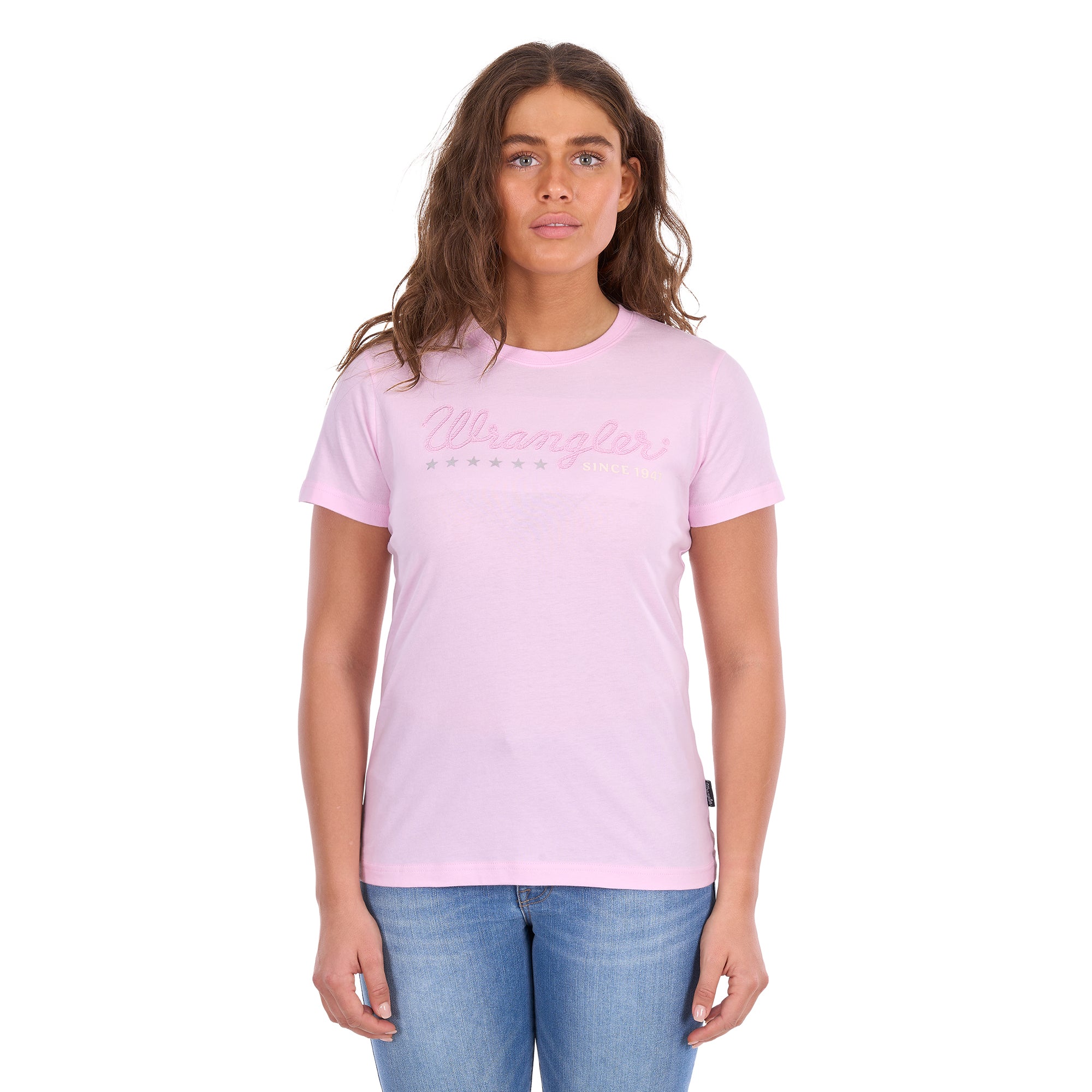 A woman modelling a pastel pink Wrangler Women's Lucy Short Sleeved T-Shirt (XCP2598633) with a pink script logo embroidered across the chest, paired with blue jeans against a plain white background.