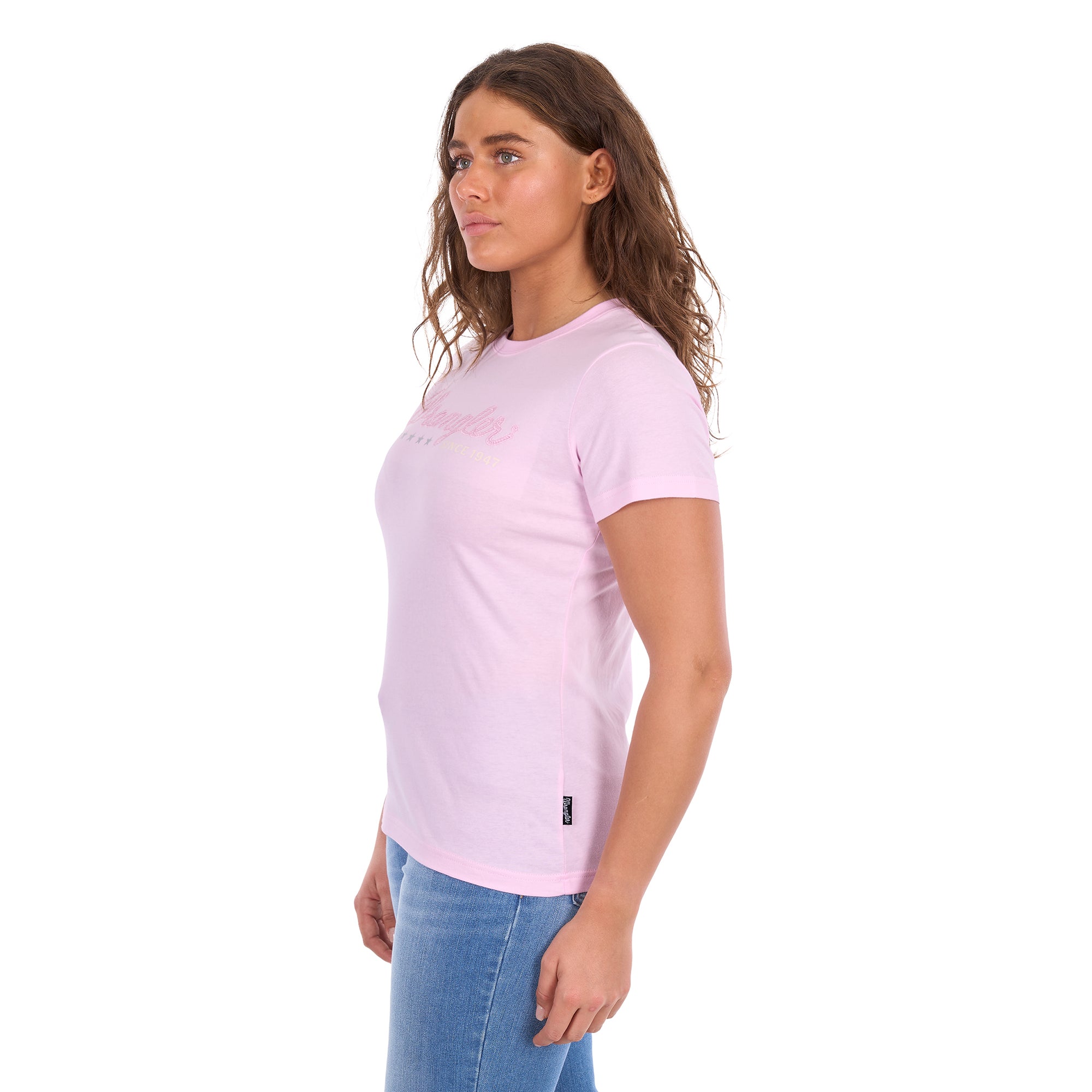 A woman in profile modelling a pastel pink Wrangler Women's Lucy Short Sleeved T-Shirt (XCP2598633) with a pink script logo embroidered across the chest, paired with blue jeans against a plain white background.