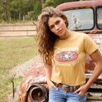 A woman with long curly brown hair stands outdoors in front of a rusted vintage truck, modelling a sunset yellow Wrangler Women's Macie Tee (X6S2598637) with a 'Wrangler Country Girl' graphic, paired with blue jeans.