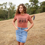 A woman stands in a dry grassy field wearing a copper Wrangler Women's Adele Tee (X6S2598644) graphic T-shirt and a faded blue Wrangler Women's Maisy Denim Skirt (XCP2409723). The shirt features a dark western-style graphic with a Wrangler logo.