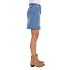 A side profile view of a faded blue Wrangler Women's Maisy Denim Skirt (XCP2409723) with decorative embroidery on the pockets, shown on a model in profile wearing tan cowboy boots against a white background.