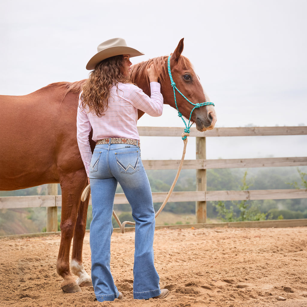 Rear view of a woman in a Wrangler Brodie Cowboy Hat (XCP1961HAT) a pink Wrangler Essie Long Sleeve Shirt (X6S2101601), and a Wrangler Blakely Belt (XCP2915BLT) wearing light blue Wrangler Women's Margo Trouser Jeans Willow (XCP2253710) with a straight-to-flare leg and decorative contrast stitching on the back pockets, stands beside a brown horse in a sandy outdoor paddock, holding the horse's reins. The horse wears a turquoise halter.