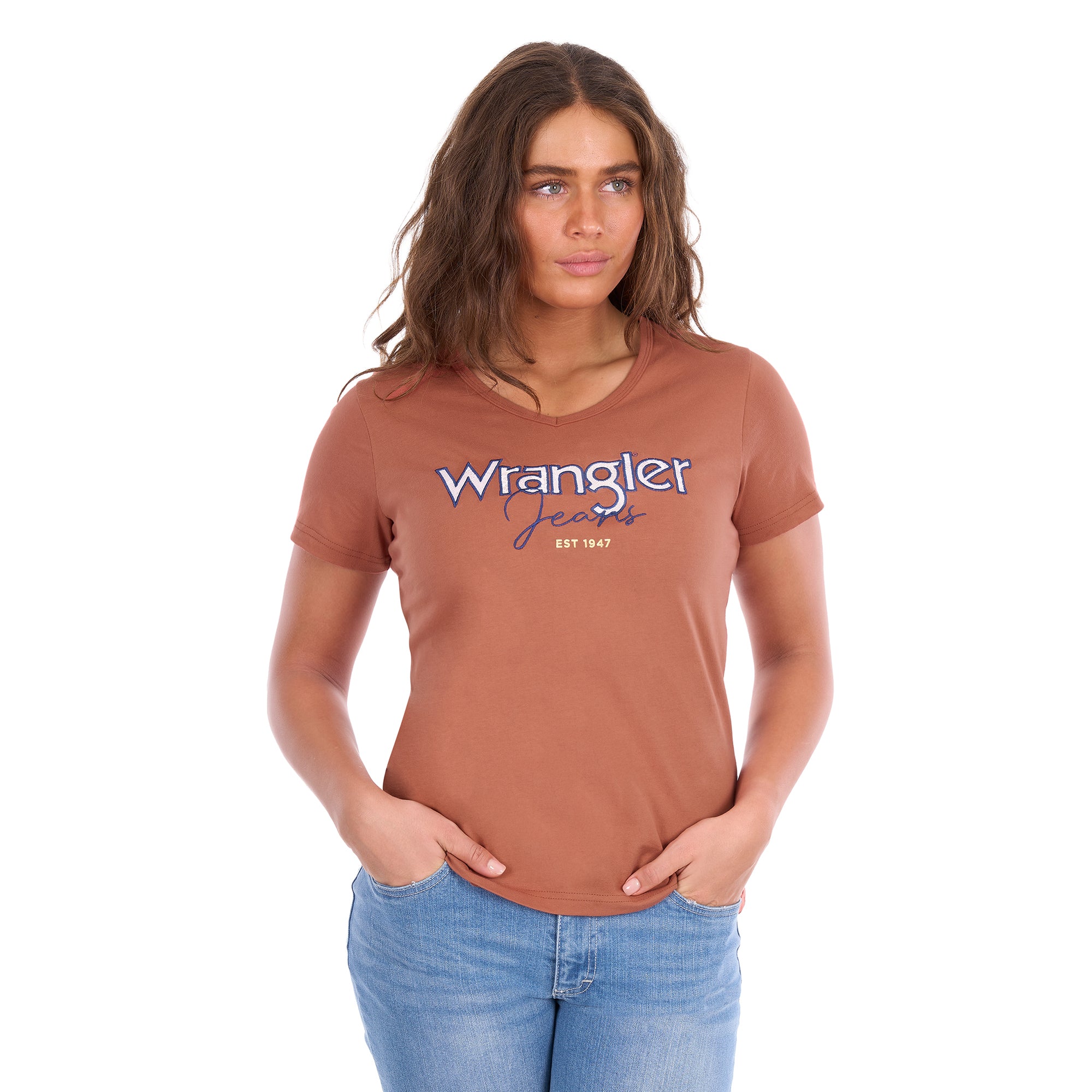 A woman modelling a Wrangler Women's Maya V-Neck S/S Tee (X6S2599640) in Copper, featuring Wrangler lace applique and embroidery with metallic detail across the chest, paired with blue jeans against a plain white background.