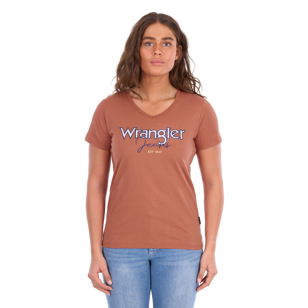 A woman modelling a Wrangler Women's Maya V-Neck S/S Tee (X6S2599640) in Copper, featuring Wrangler lace applique and embroidery with metallic detail across the chest, paired with blue jeans against a plain white background.