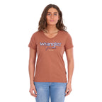 A woman modelling a Wrangler Women's Maya V-Neck S/S Tee (X6S2599640) in Copper, featuring Wrangler lace applique and embroidery with metallic detail across the chest, paired with blue jeans against a plain white background.