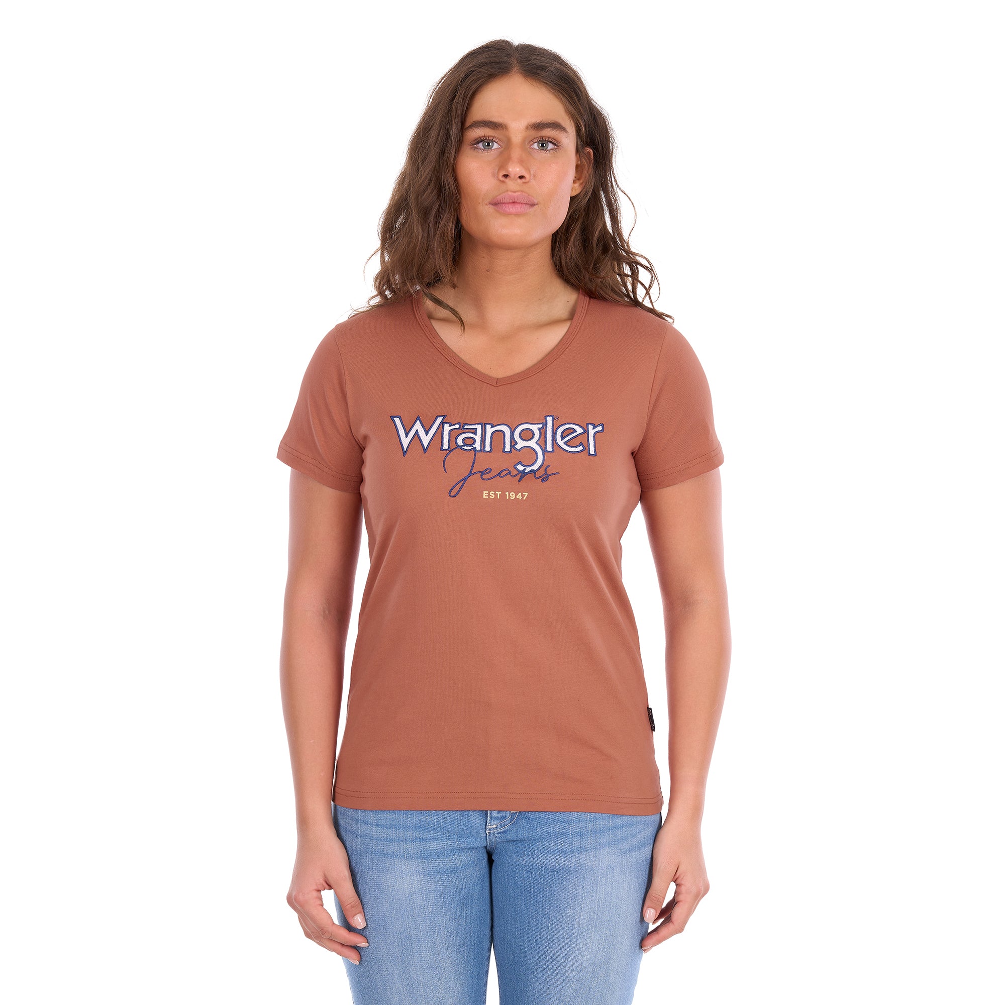 A woman modelling a Wrangler Women's Maya V-Neck S/S Tee (X6S2599640) in Copper, featuring Wrangler lace applique and embroidery with metallic detail across the chest, paired with blue jeans against a plain white background.