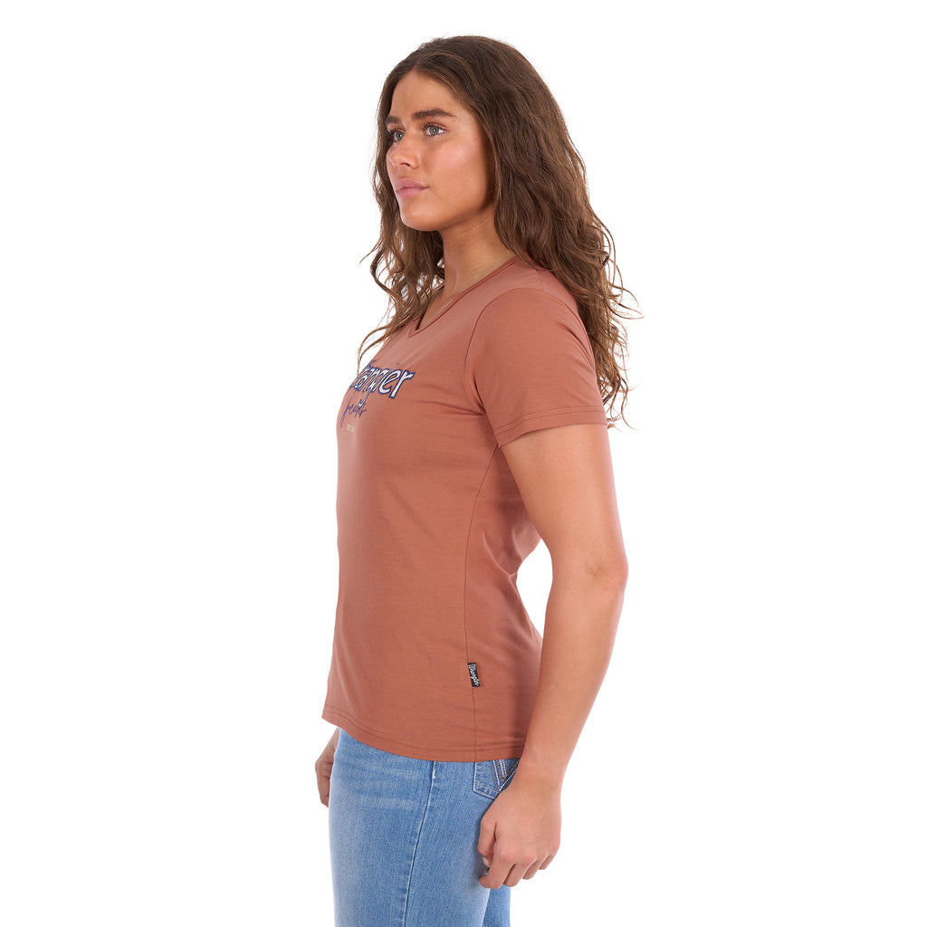 A woman viewed in side profile modelling a Wrangler Women's Maya V-Neck S/S Tee (X6S2599640) in Copper, featuring Wrangler lace applique and embroidery with metallic detail across the chest, paired with blue jeans against a plain white background.