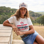 A woman sitting outdoors on a wooden bench modelling a Wrangler Women's Nora Short Sleeve Tee (XCP2598636) in cream, featuring a red, white, and navy blue graphic logo on the chest, paired with a Wrangler Women's Maisy Denim Skirt (XCP2409723) a Wrangler Siara Belt (X6S2916BLT) and a matching Wrangler Nora High Profile Full Back Cap (XCP2947CAP), with a blurred natural landscape in the background.