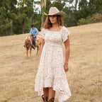 A woman modelling a cream Wrangler Women's Renee Short Sleeved Dress (X6S2407621) with a square neckline and a delicate western floral print on a light cream background, paired with brown ankle boots and a Silverton Wrangler Brodie Hat (XCP1961HAT) in a paddock with a horse and rider and trees in the background.