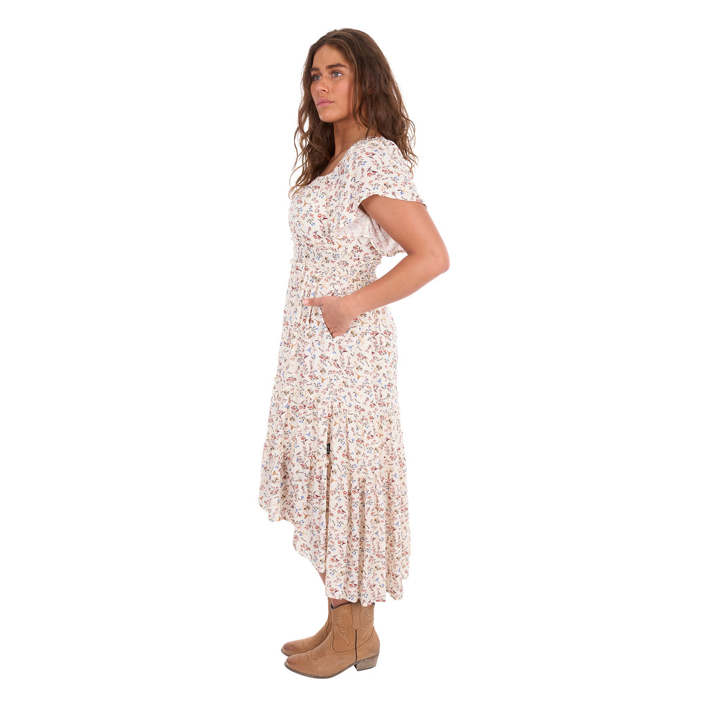 A woman modelling a cream Wrangler Women's Renee Short Sleeved Dress (X6S2407621) with a square neckline and a delicate western floral print on a light cream background, paired with brown ankle boots, set against a plain white background, photographed from the side.