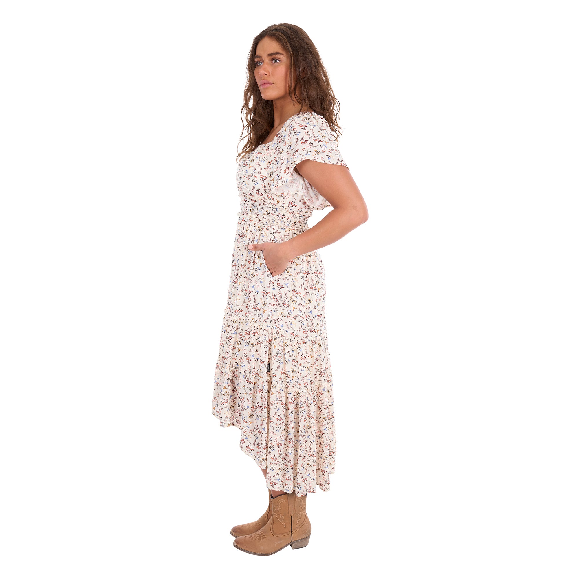 A woman modelling a cream Wrangler Women's Renee Short Sleeved Dress (X6S2407621) with a square neckline and a delicate western floral print on a light cream background, paired with brown ankle boots, set against a plain white background, photographed from the side.