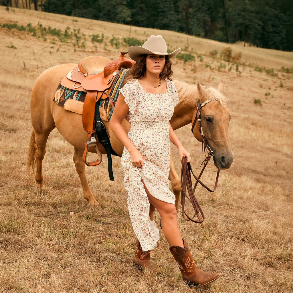 A woman modelling a cream Wrangler Women's Renee Short Sleeved Dress (X6S2407621) with a square neckline and a delicate western floral print on a light cream background, paired with brown ankle boots and a Silverton Wrangler Brodie Hat (XCP1961HAT) leading a horse in western tack in a dry paddock.
