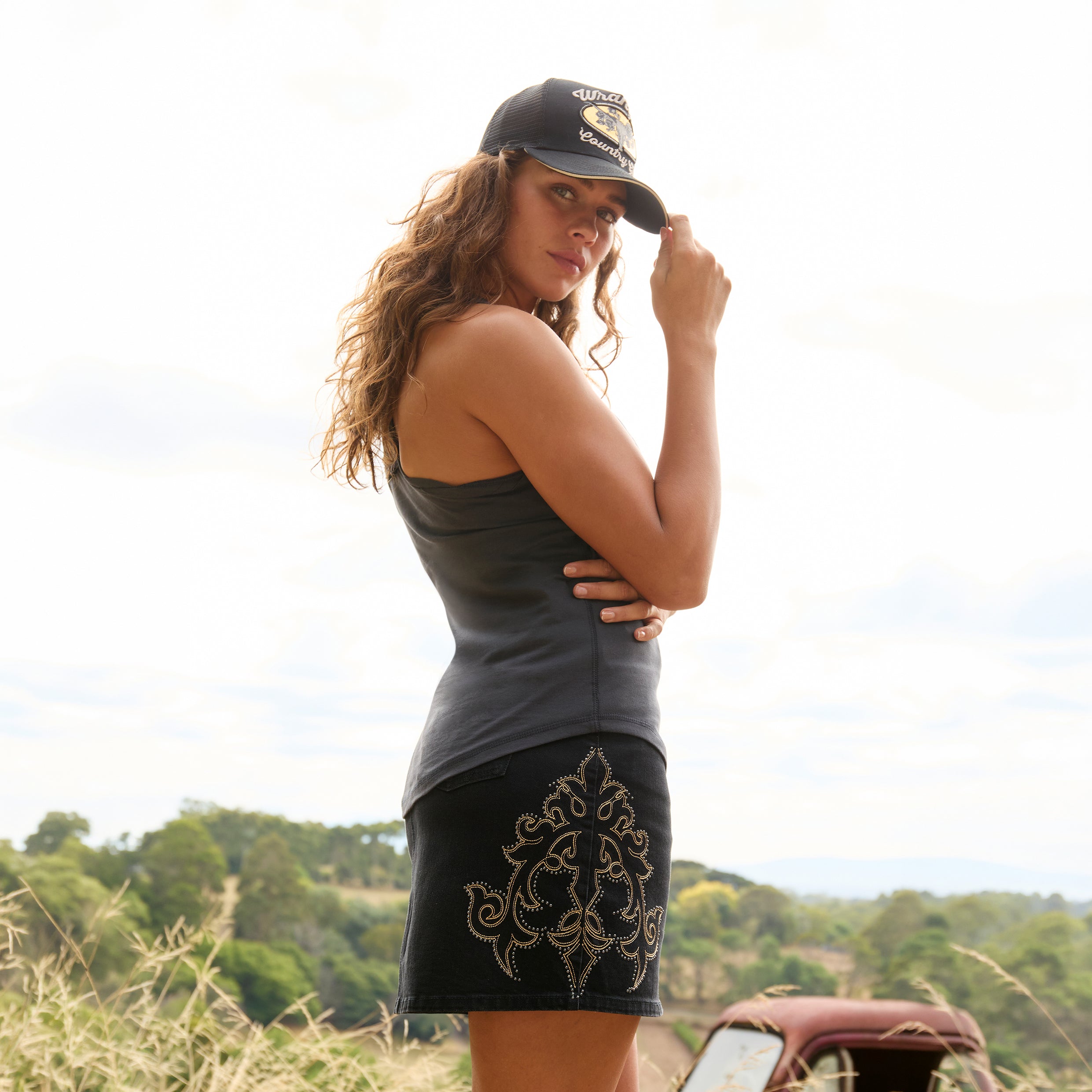 A woman modelling a black Wrangler Women's Rock Denim Mini Skirt (X6S2409725) with silver embroidered floral details on the sides, a Wrangler Jasmine Tank Top, and a Jasmine Trucker Cap, photographed outdoors.