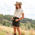 A woman wearing a cream Wrangler Women's Cindy Crop Short Sleeved Tee (XCP2596647) with scripted 'Wrangler' printed across the chest, a black Wrangler Women's Rock Denim Skirt (X6S2409725) with studs and gold embroidery, a Wrangler Kenzley Belt (X6S2917BLT) and a Wrangler Brodie Hat (XCP1961HAT) Silverton cowboy hat, standing outdoors in a grassy field with trees in the background.
