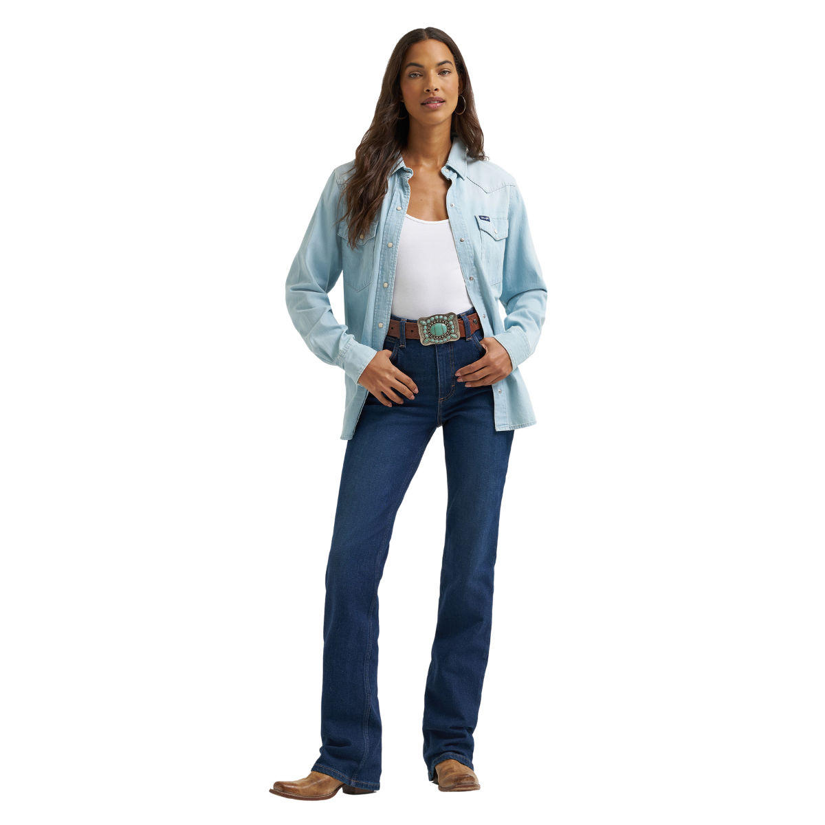 A woman modelling a pair of Kayla blue Wrangler Women's Rodeo Bootcut Jeans (112367606) paired with a white top, an open blue shirt, a western belt and brown western boots, on a white studio background.