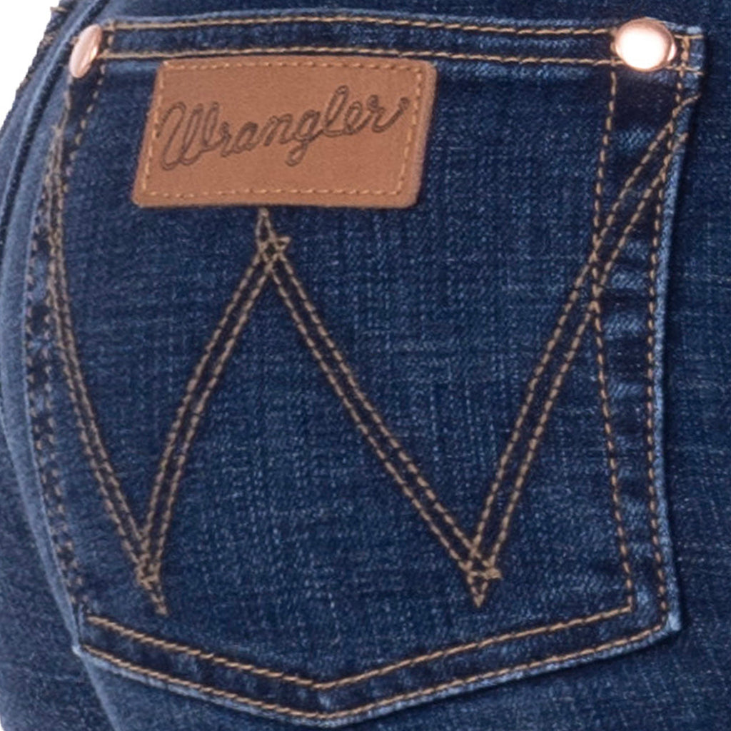 Close-up of the back pocket of a person wearing old indigo blue Wrangler Women's Sierra Skinny Jeans (XCP2238861), showing the brown leather brand patch, contrast stitching, and metal rivets on a white background.
