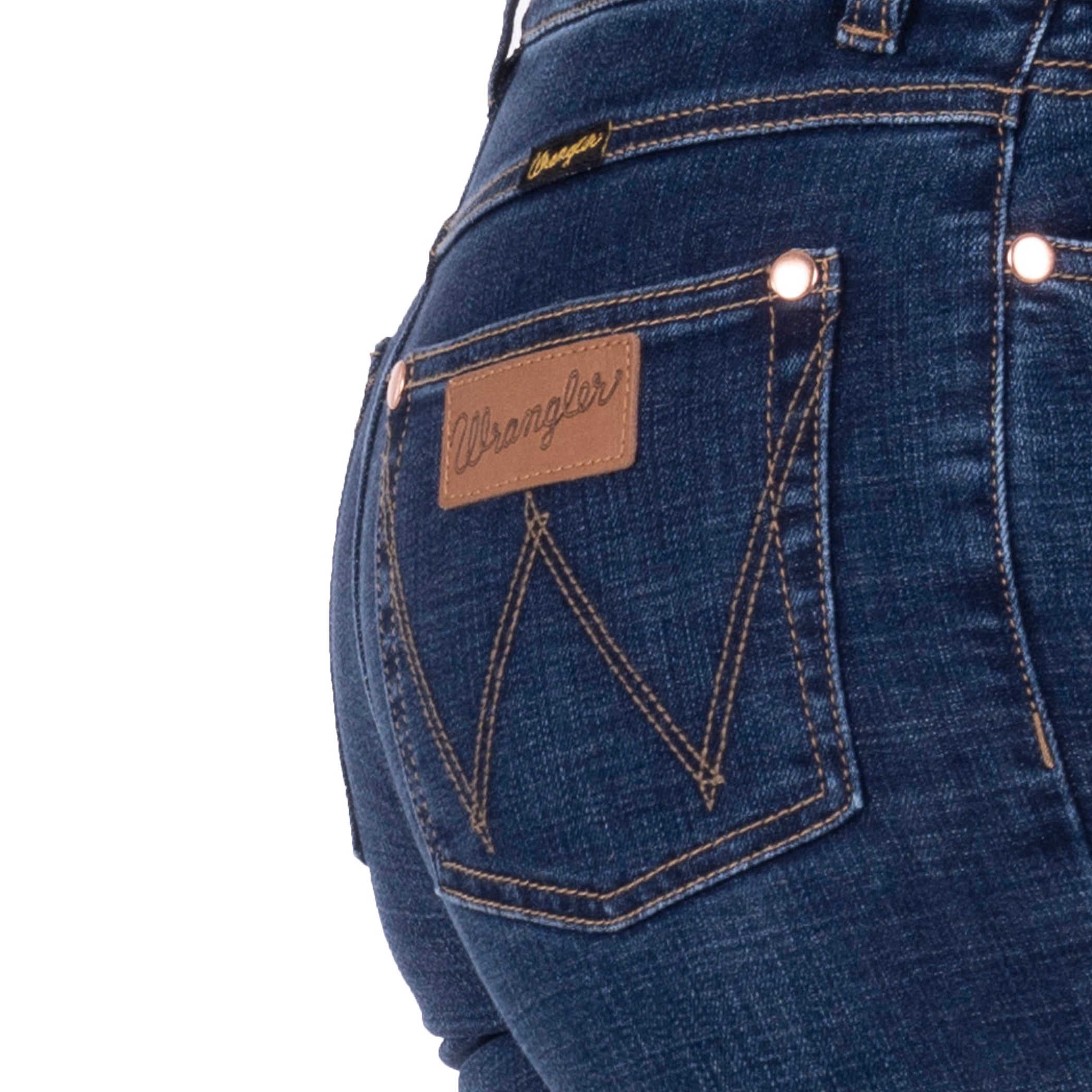Close-up of the back pocket of a person wearing old indigo blue Wrangler Women's Sierra Skinny Jeans (XCP2238861), showing the brown leather brand patch, contrast stitching, and metal rivets on a white background.