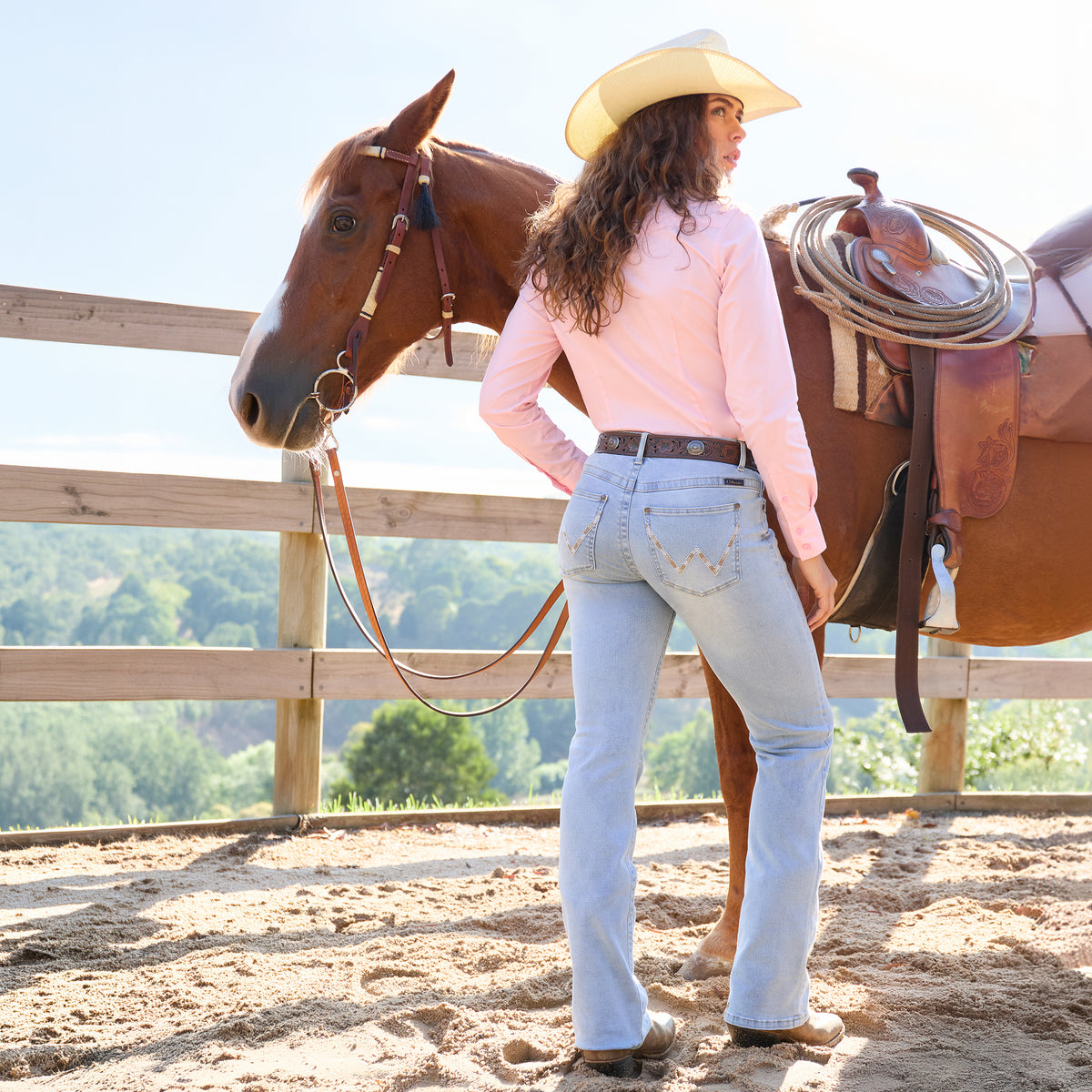 A woman in a blush pink Wrangler Womens Hazel Logo L/S Shirt (XCP2111608), sky light blue Wrangler Women's Verity Bootcut Jeans - Willow (X6S2251722) featuring signature Wrangler W stitching on the pockets, and a Wrangler Jhonson Bangora Cowboy Hat (X4Y2025JHO) stands beside a horse in a sunny outdoor paddock, viewed from behind.
