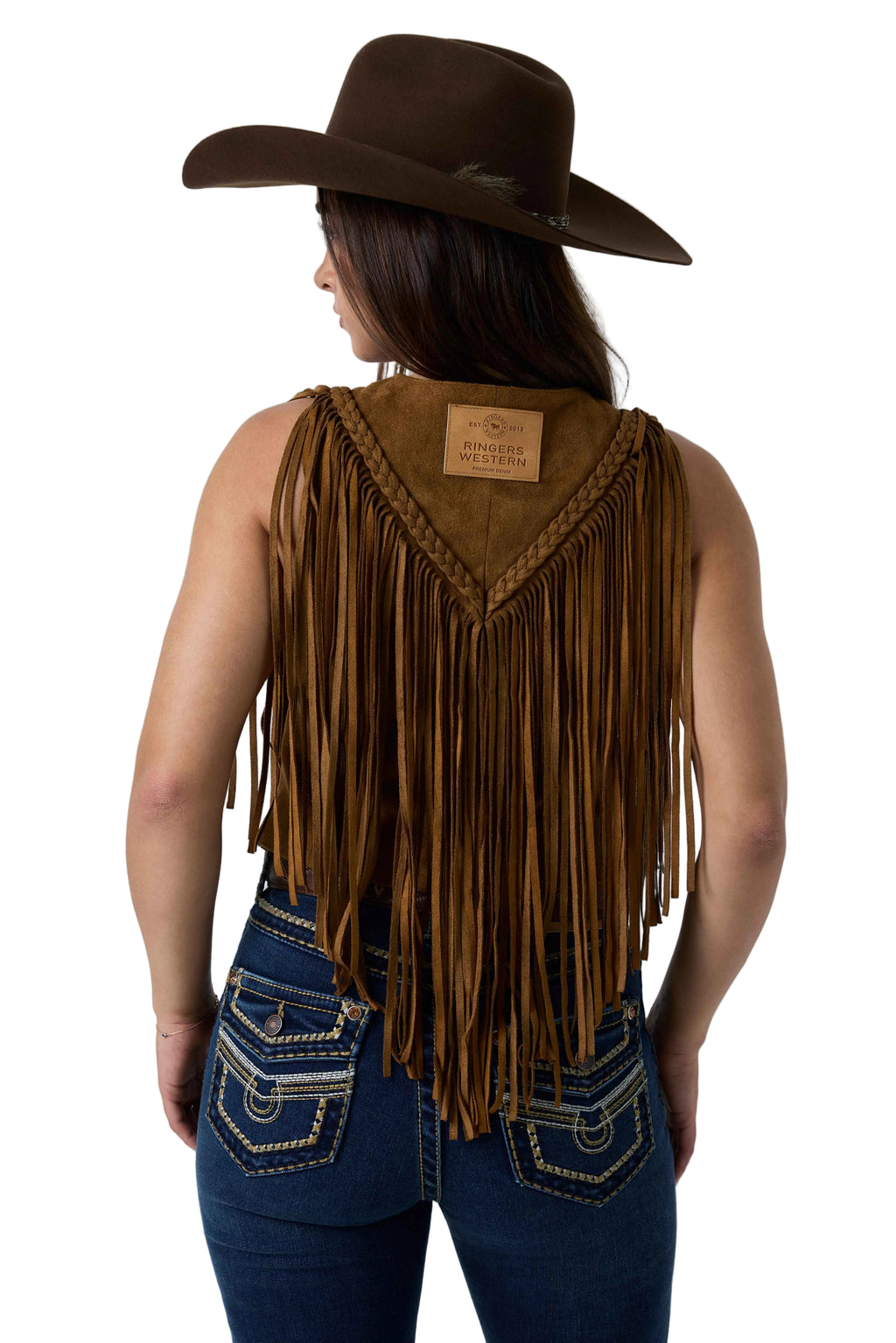 A woman shown from the back wearing a brown suede fringe vest with braided trim, a matching brown cowboy hat, and blue jeans against a white studio background.