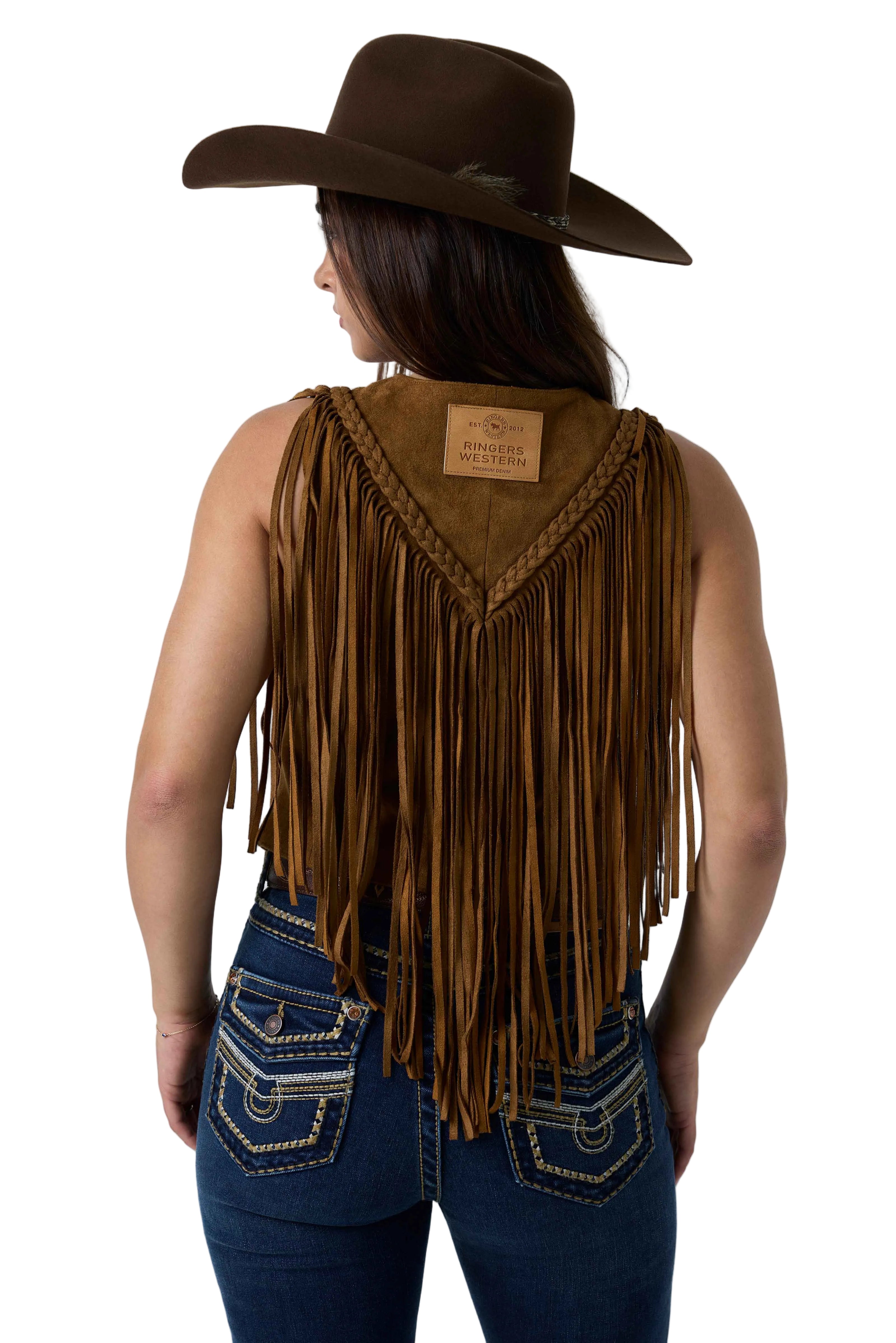 A woman shown from the back wearing a brown suede fringe vest with braided trim, a matching brown cowboy hat, and blue jeans against a white studio background.