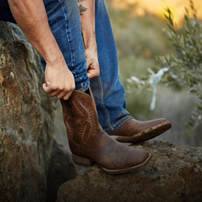 A person wearing blue jeans pulls on a brown cowboy boot while standing on a rock outdoors, with plants and rocks visible in the background.