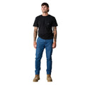 A man with short brown hair and arm tattoos stands facing forward, wearing a black t-shirt, x/dmg Mens Denim Work Pants (x01/DENIM) 90's Blue, and tan lace-up boots against a plain white background.